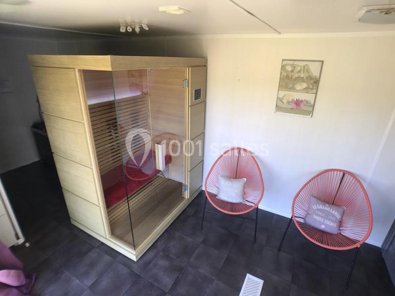 Cabine de sauna en bois avec porte vitrée, deux fauteuils orange et un tableau décoratif sur un mur blanc.