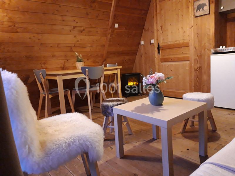 Intérieur d'un chalet en bois avec table, chaises recouvertes de fourrure, poêle allumé et vase de fleurs sur une table…