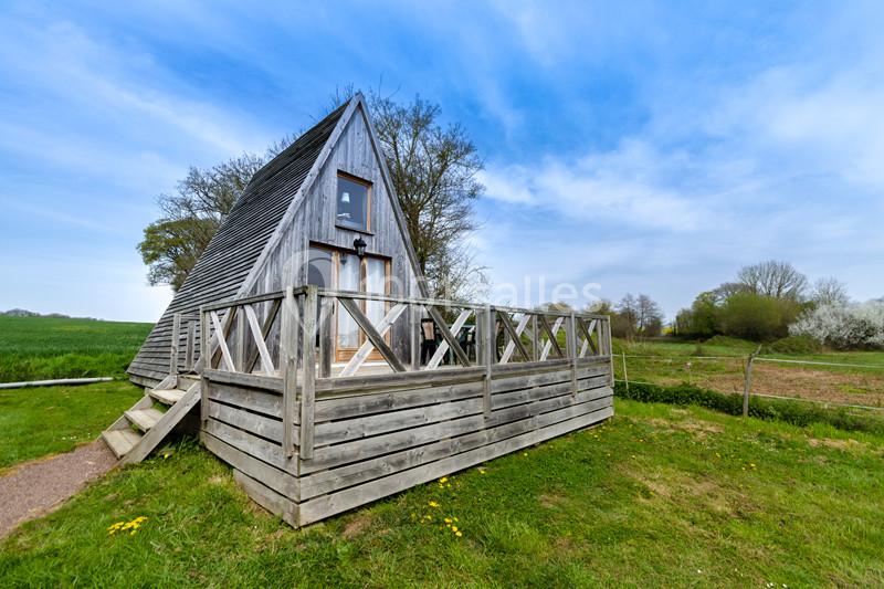 Cabane en bois en forme de A avec terrasse, située dans un paysage rural verdoyant sous un ciel bleu.