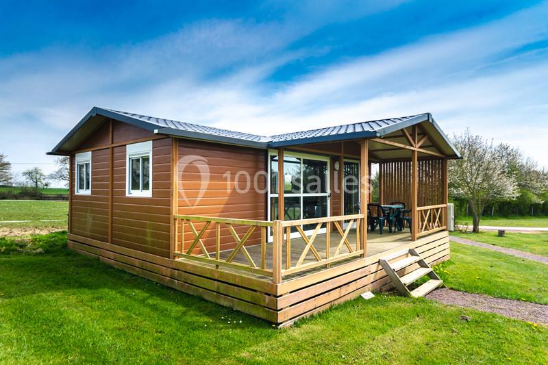 Chalet en bois avec terrasse, situé sur une pelouse verte, entouré d'arbres sous un ciel bleu.