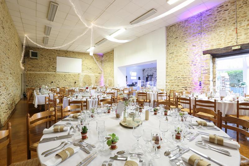 Salle de réception décorée avec des tables rondes dressées, chaises en bois et guirlandes lumineuses au plafond.