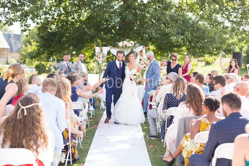 Un couple de mariés marche dans une allée extérieure bordée d'invités assis, sous un arbre décoré de fleurs.