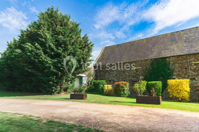 Vue d'un bâtiment en pierre entouré d'arbustes colorés, avec un grand arbre et un chemin en gravier au premier plan.