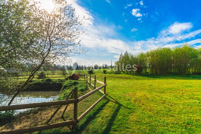 Paysage champêtre avec une clôture en bois, un étang à gauche et une prairie verdoyante sous un ciel bleu.