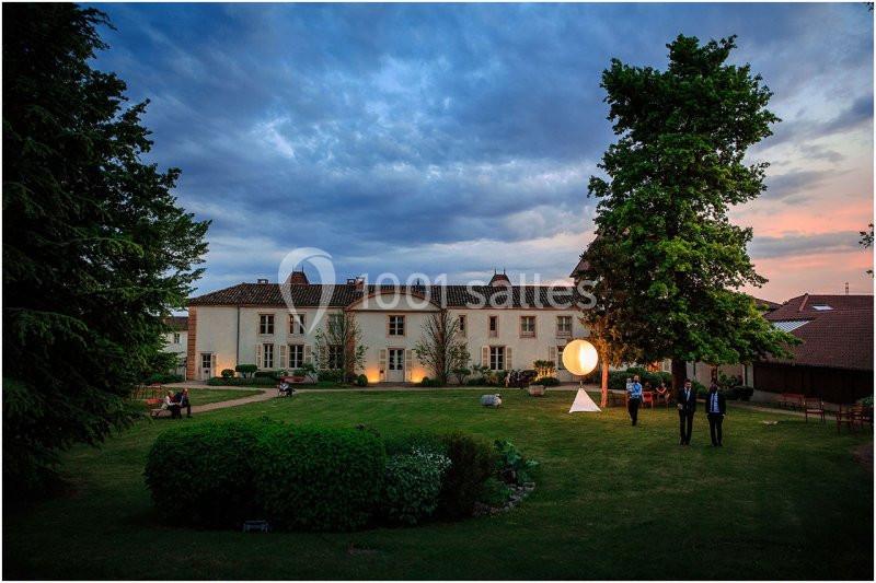 Vue d'un grand bâtiment ancien entouré d'un jardin verdoyant au crépuscule, avec quelques personnes discutant.