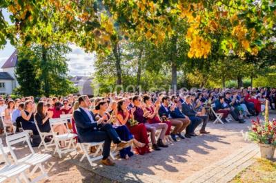 Des invités assis en plein air applaudissent lors d'une cérémonie sous des arbres aux feuilles partiellement jaunies.