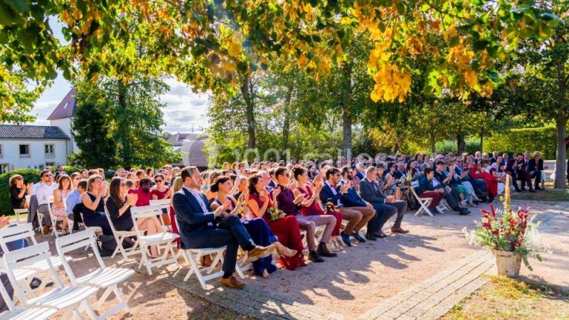 Des invités assis en plein air applaudissent lors d'une cérémonie sous des arbres aux feuilles partiellement jaunies.