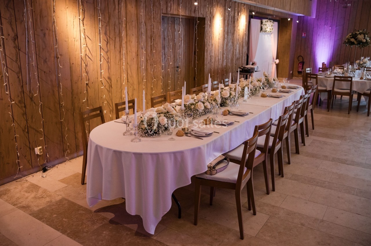 Grande table de réception décorée de fleurs blanches, bougies et nappes blanches, dans une salle en bois éclairée.