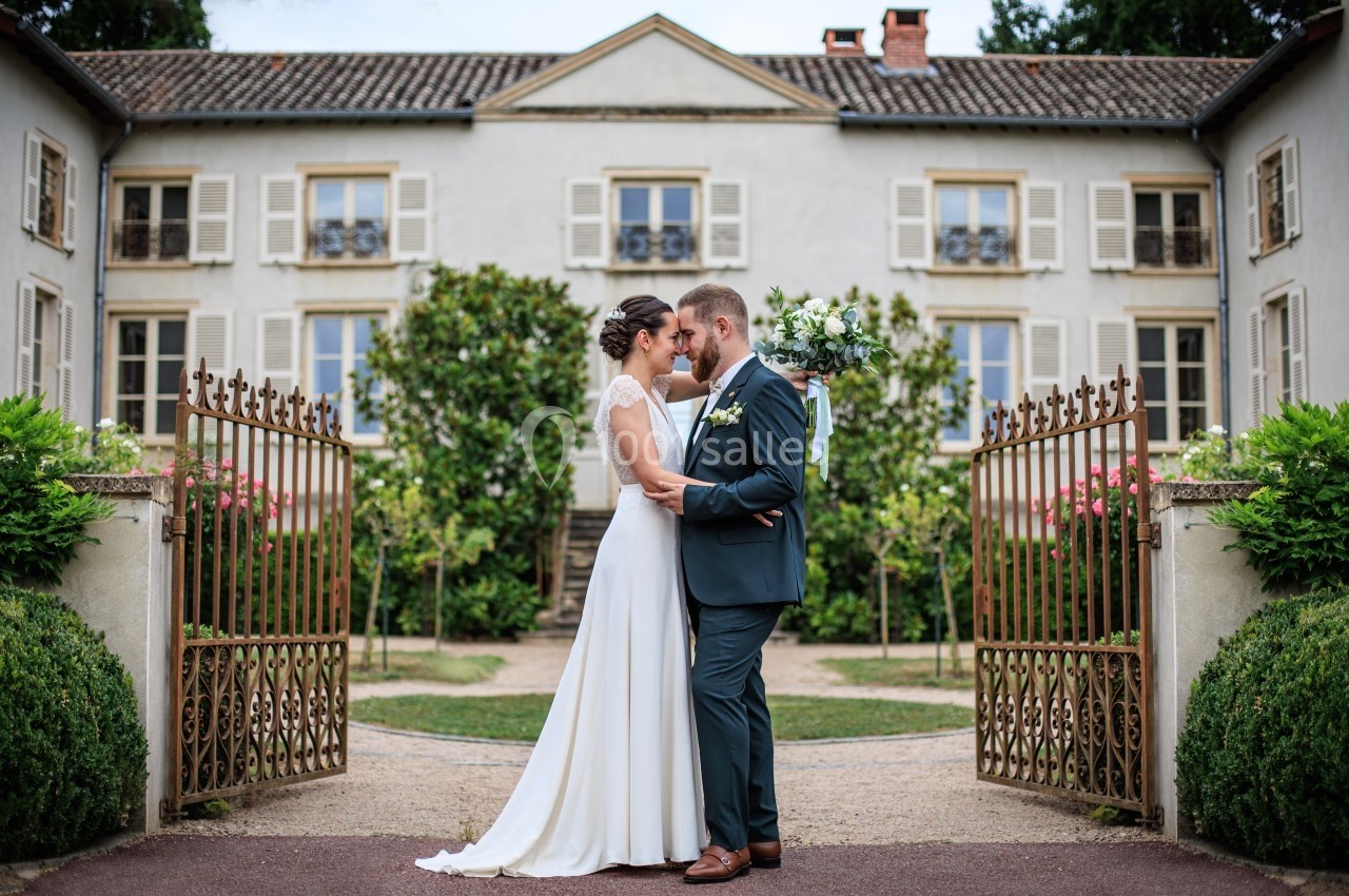 Un couple de mariés se tient face à face devant un portail en fer forgé et une maison élégante.