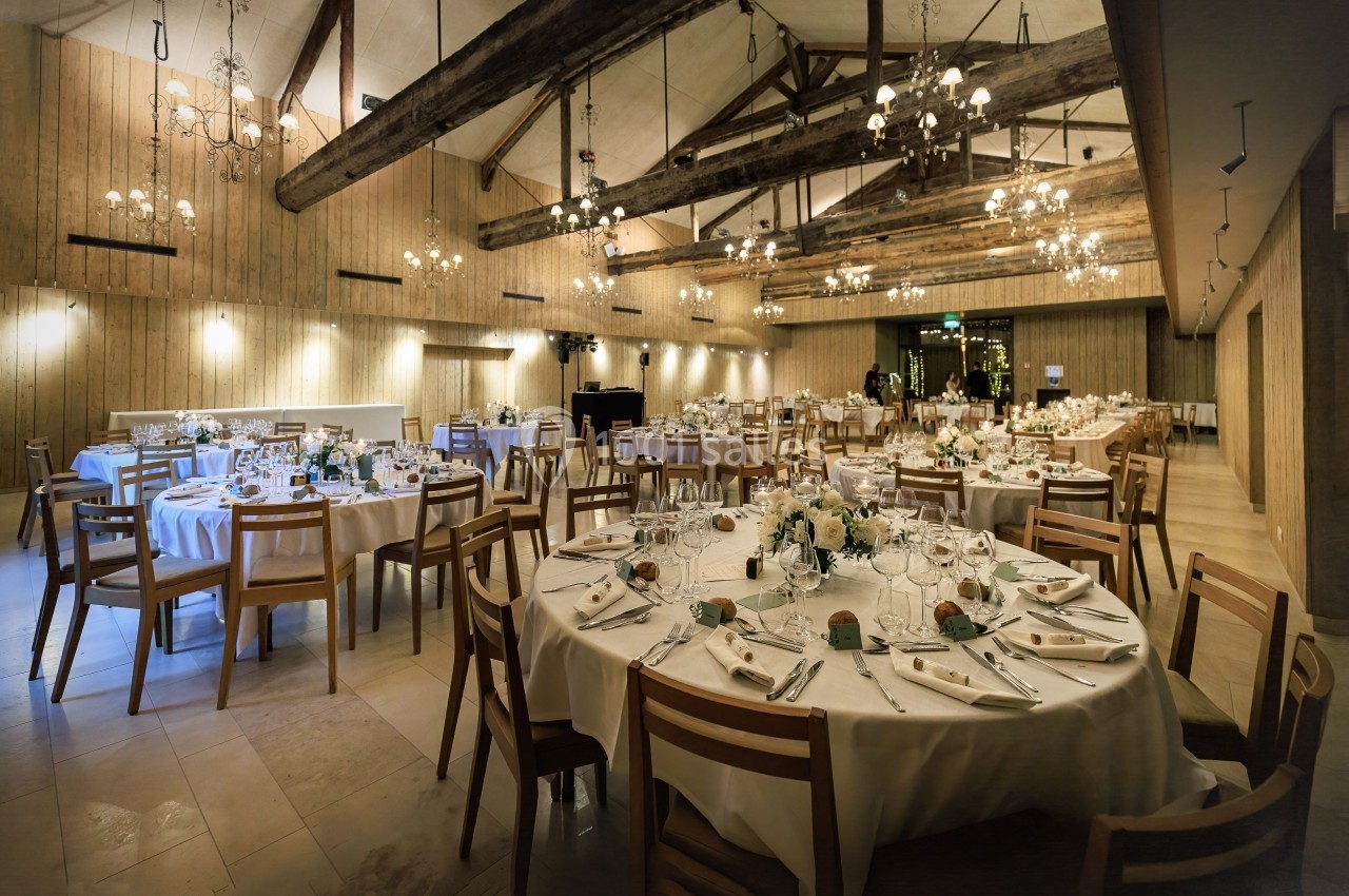 Salle de réception avec tables rondes dressées, décorations florales et poutres apparentes au plafond.