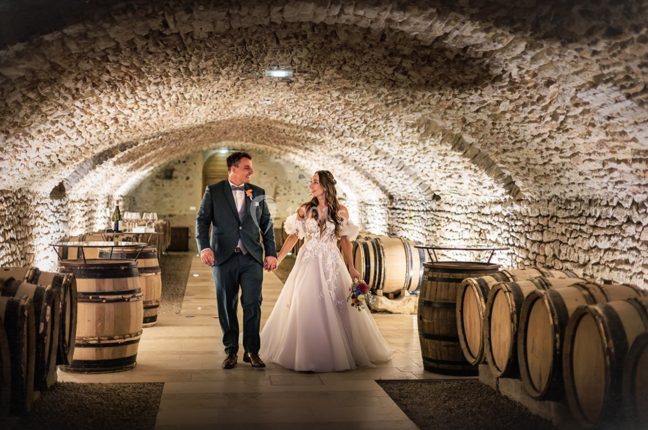 Un couple en tenue de mariage marche main dans la main dans une cave voûtée entourée de tonneaux en bois.