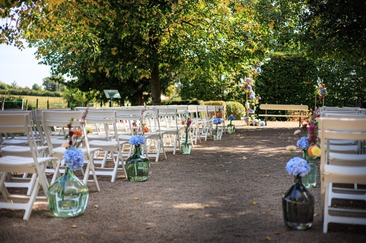 Chaises blanches alignées en extérieur, décorées de fleurs, formant une allée sous des arbres pour une cérémonie.