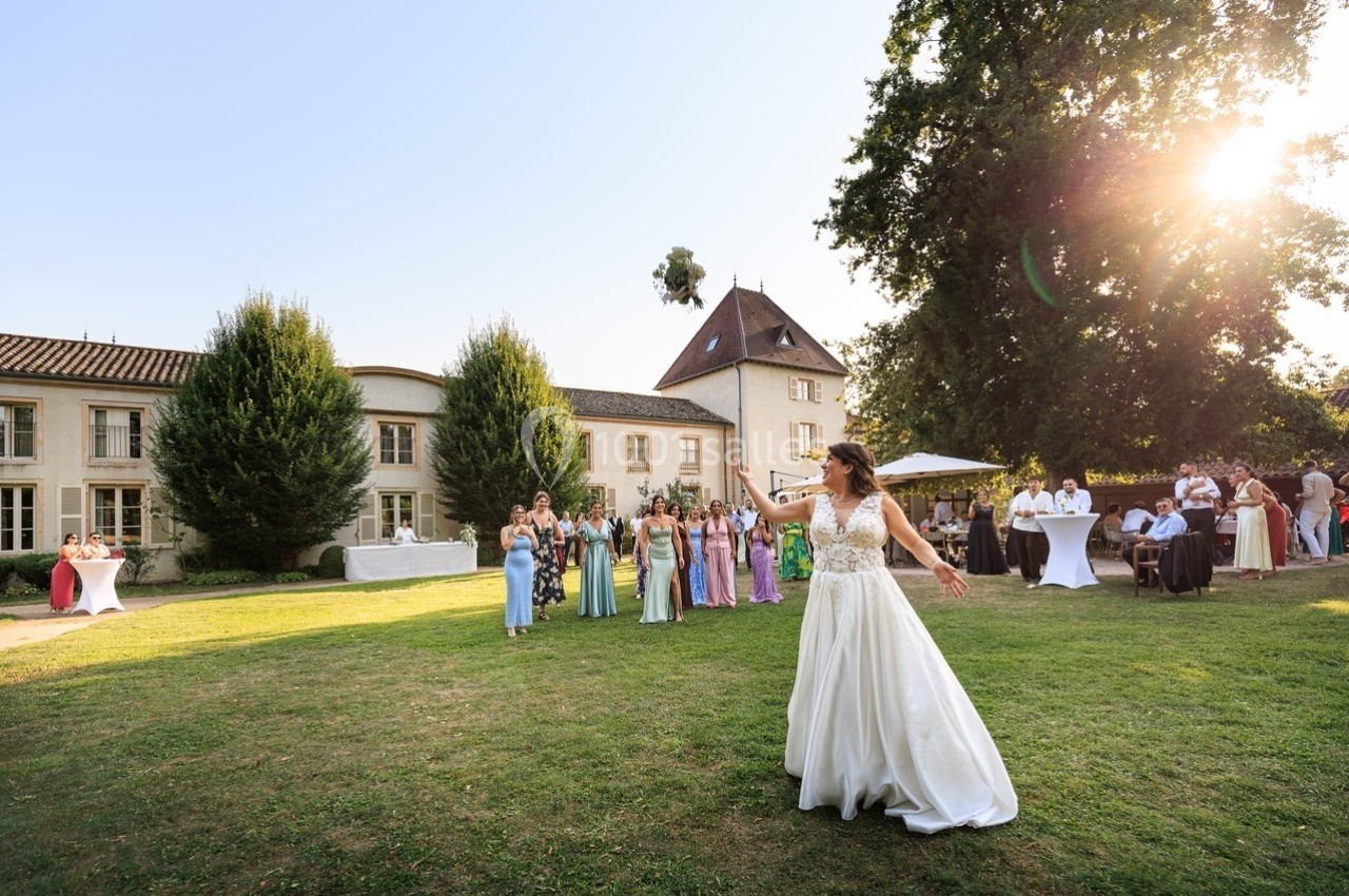 Une mariée lance son bouquet devant des invitées rassemblées dans le jardin d'un domaine sous un soleil couchant.