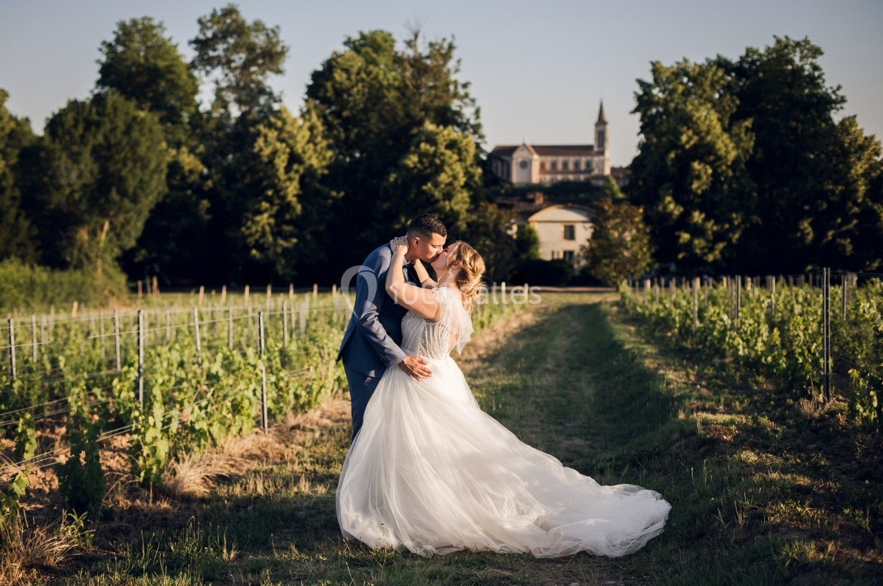 Un couple en tenue de mariage s'embrasse dans un vignoble au coucher du soleil, avec une église en arrière-plan.