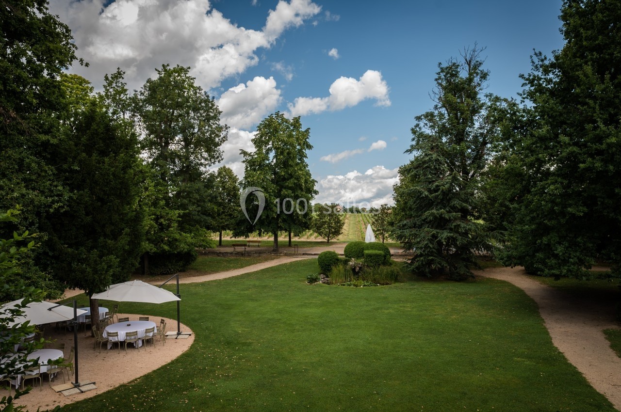 Jardin verdoyant avec pelouse, arbres, chemins en gravier et tables sous parasols par une journée ensoleillée.