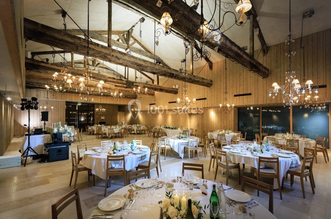 Salle de réception spacieuse avec poutres apparentes, tables rondes dressées et éclairage chaleureux.