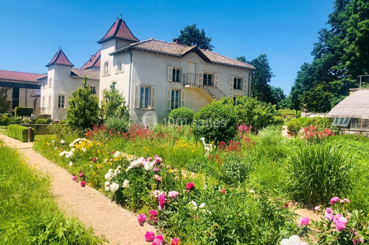 Jardin fleuri avec des allées bordées de fleurs colorées devant une maison ancienne à toits en tuiles rouges.
