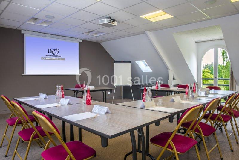 Salle de réunion lumineuse avec tables disposées en U, chaises rouges, écran de projection et vue sur l'extérieur.