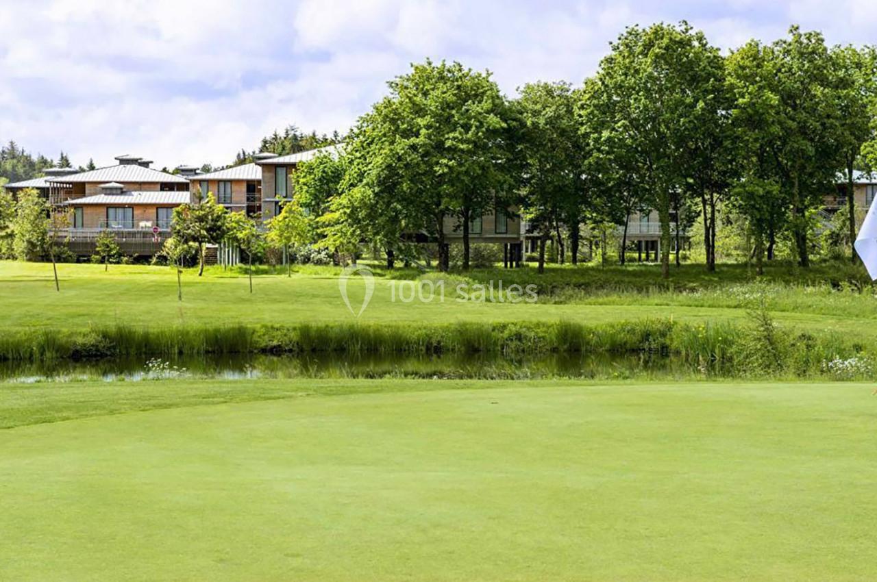 Vue d'un terrain de golf verdoyant avec un étang, des arbres et des bâtiments modernes en arrière-plan.