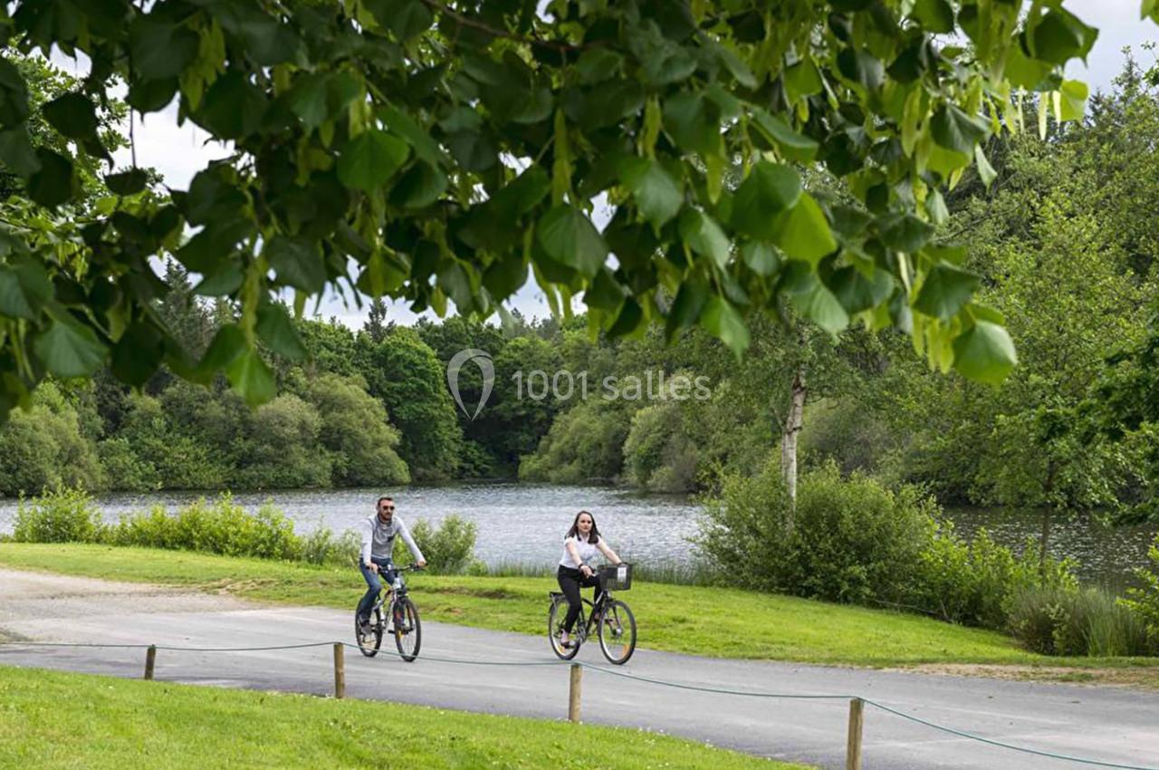Deux personnes font du vélo sur un chemin bordé d'arbres, avec un lac et une végétation dense en arrière-plan.