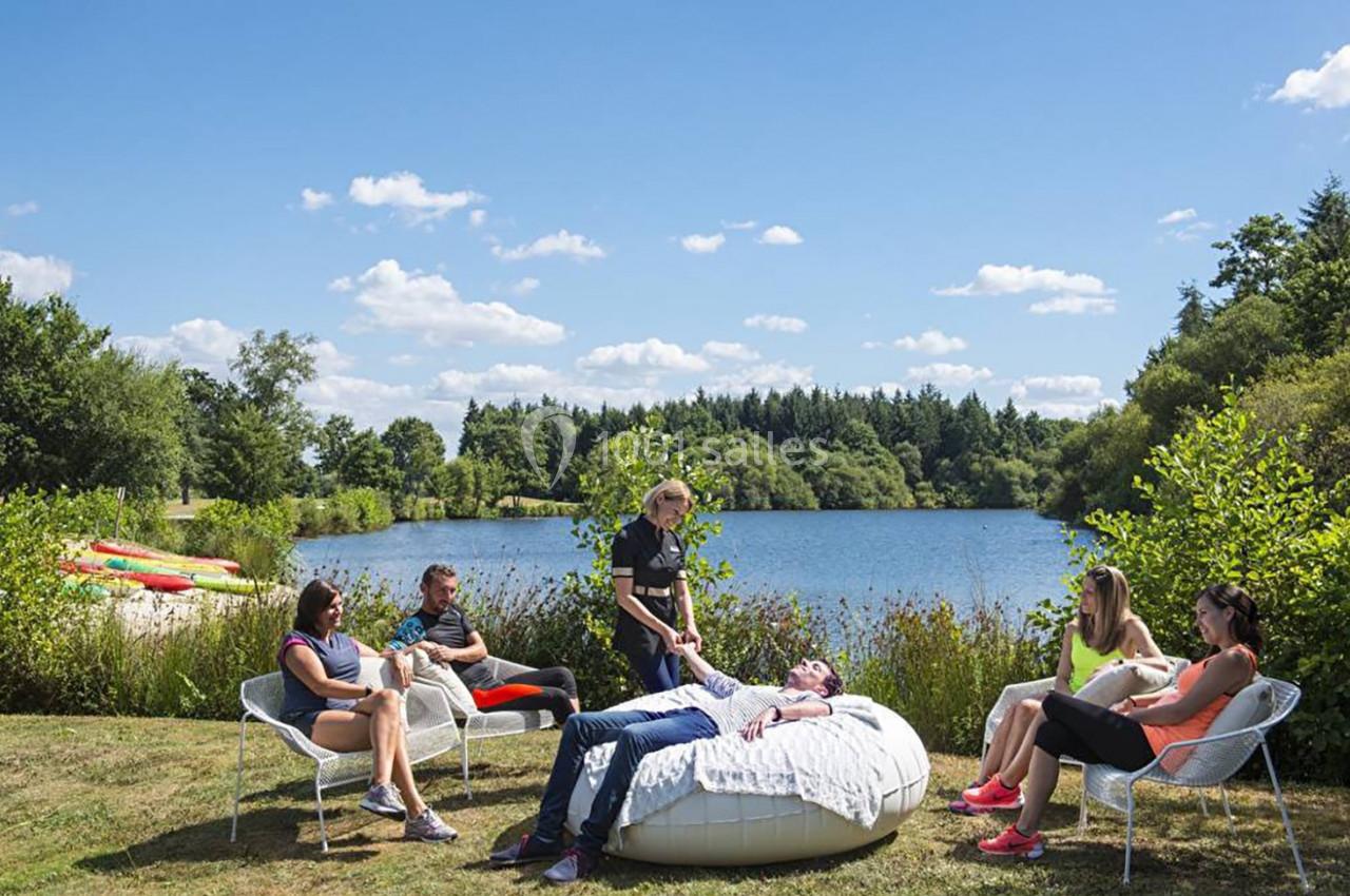 Un groupe de personnes assises près d'un lac, entouré de verdure, avec une personne allongée sur un pouf.