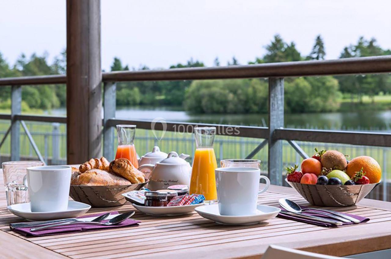 Petit-déjeuner servi en terrasse avec croissants, fruits, jus d'orange et vue sur un lac entouré de verdure.