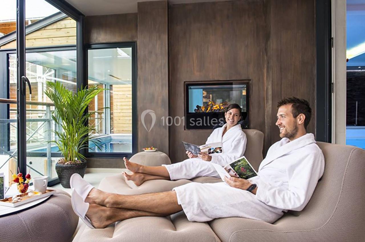 Deux personnes en peignoir se détendent dans un salon lumineux près d'une cheminée, avec des brochures en main.