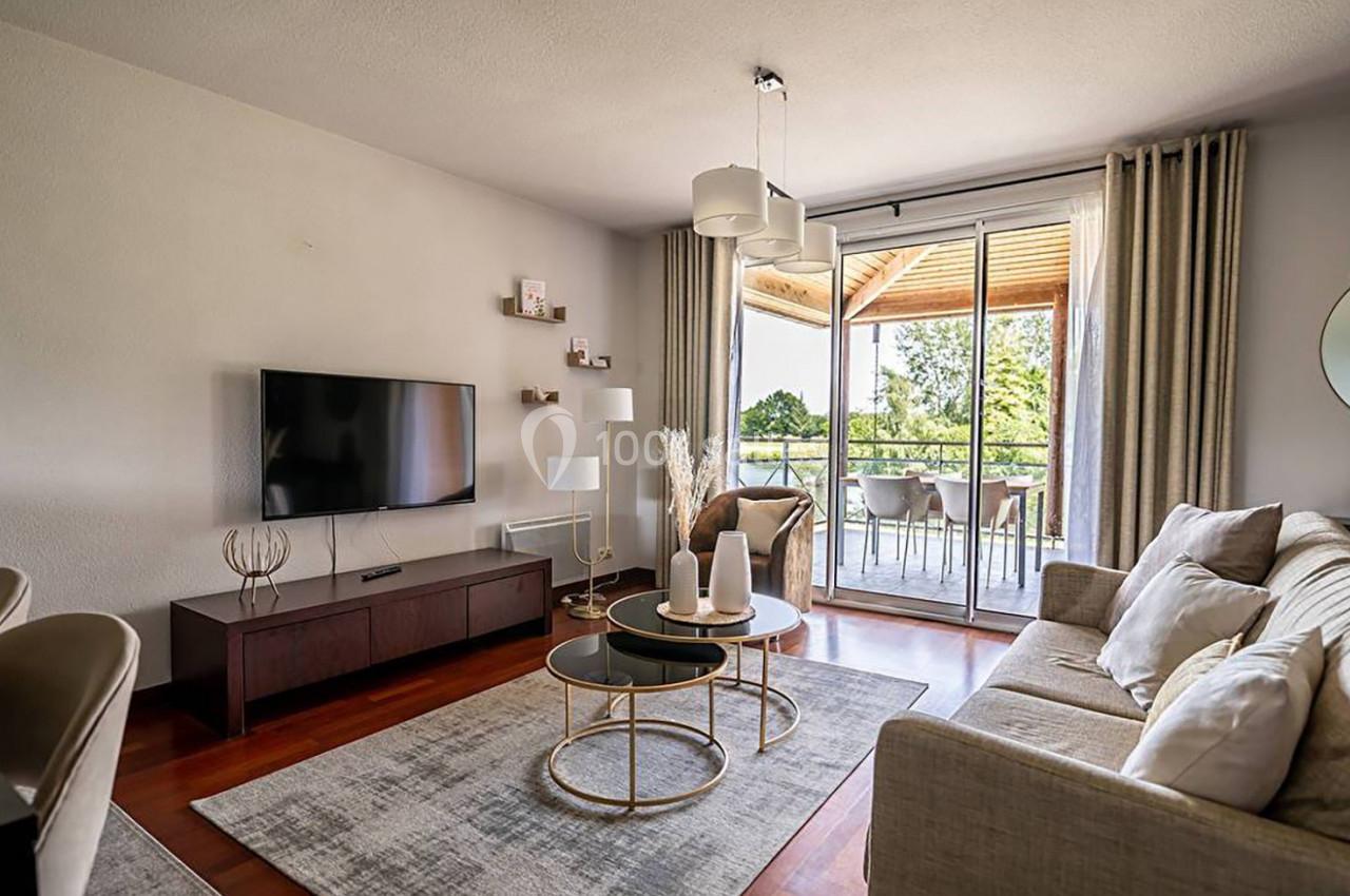 Salon lumineux avec canapé beige, table basse ronde, téléviseur mural et baie vitrée donnant sur une terrasse avec vue sur…