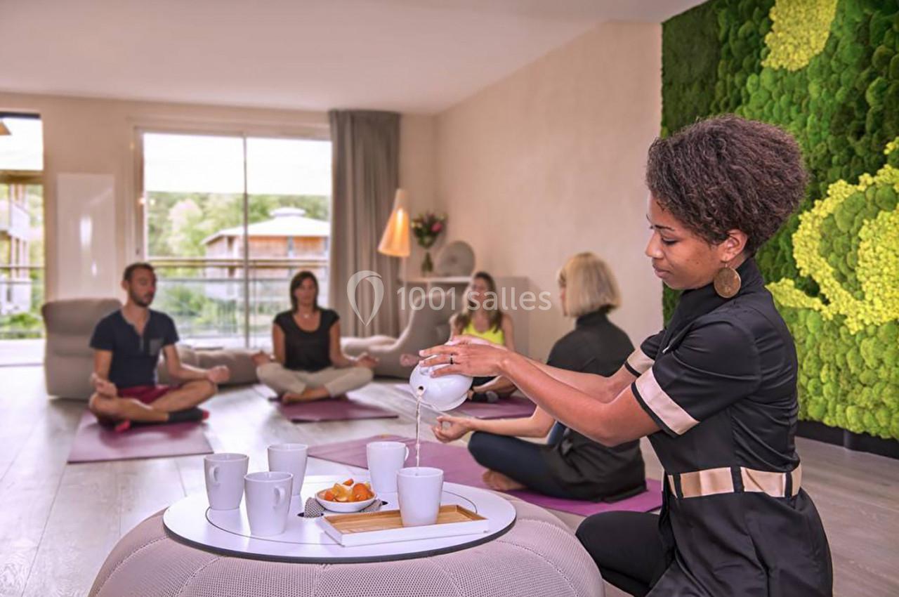 Une femme sert du thé dans une salle lumineuse où des personnes pratiquent la méditation sur des tapis.