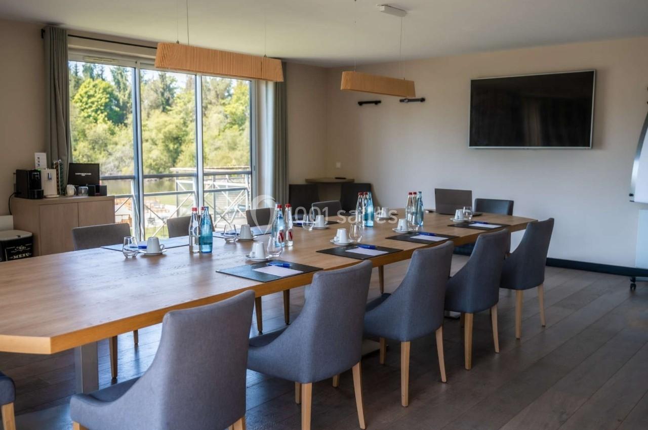 Salle de réunion lumineuse avec une grande table en bois, chaises grises, écran mural et vue sur l'extérieur verdoyant.
