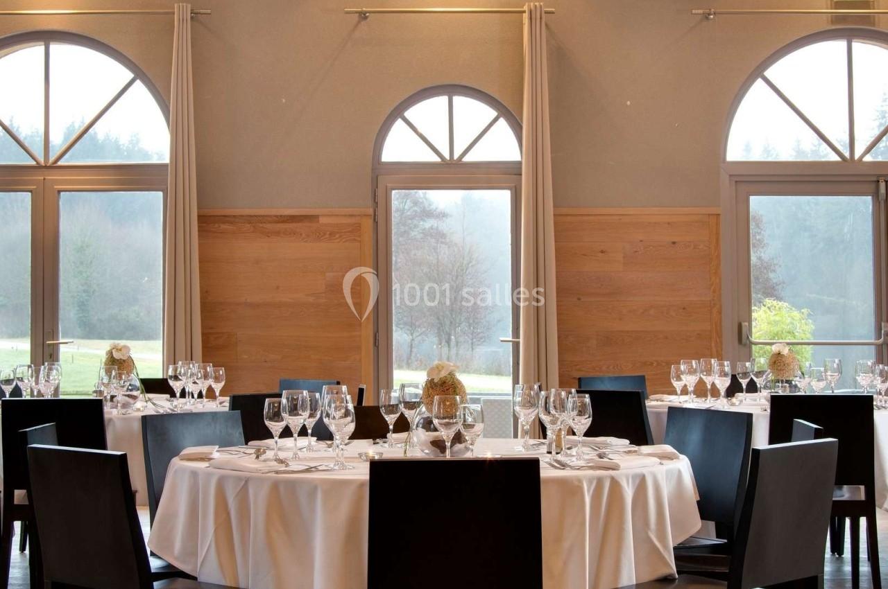Salle de réception lumineuse avec tables rondes dressées, nappes blanches, chaises noires et grandes fenêtres en arche.