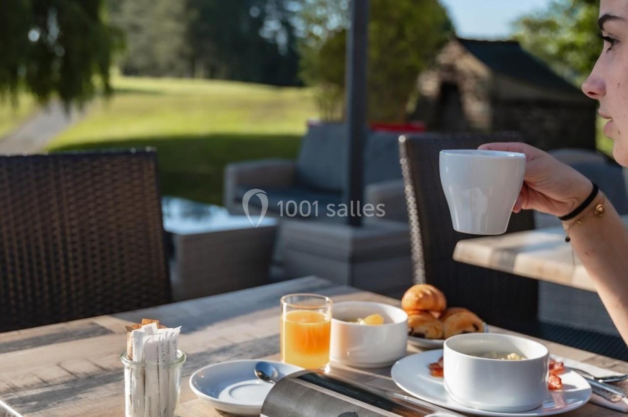 Une personne prend un café en terrasse avec un petit-déjeuner composé de viennoiseries, jus d'orange et confiture.