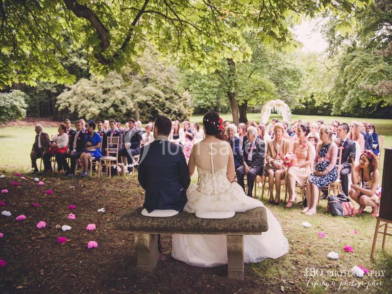Un couple assis sur un banc en pierre lors d'une cérémonie de mariage en plein air, entouré d'invités.