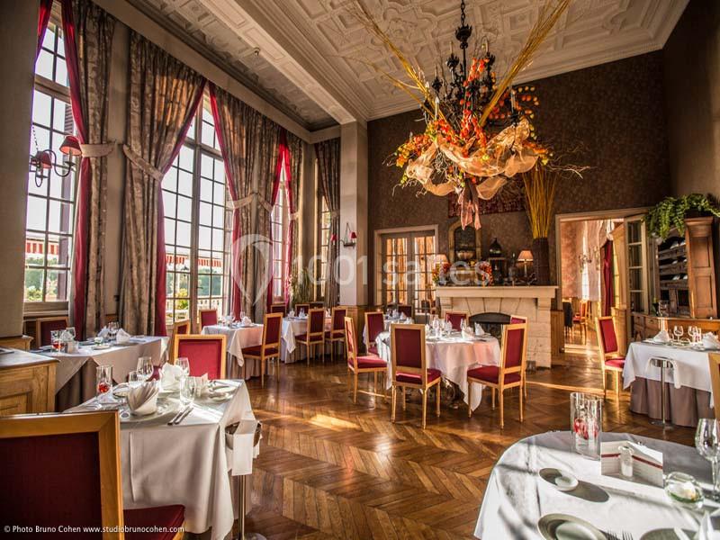 Salle de restaurant lumineuse avec grandes fenêtres, tables dressées et décoration florale suspendue au plafond.