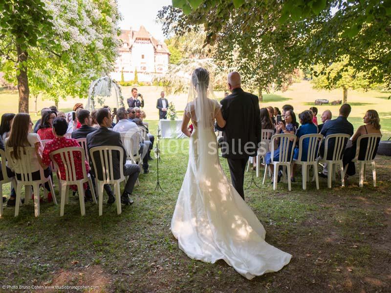 Une mariée et son accompagnateur avancent vers une cérémonie de mariage en plein air, entourés d'invités assis.