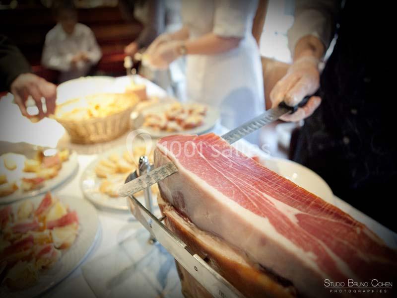 Un jambon entier est tranché à la main sur une table garnie de plateaux de charcuterie et de pain.