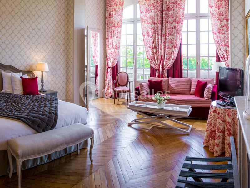 Chambre lumineuse avec parquet en bois, lit double, canapé rouge, table basse et grandes fenêtres avec rideaux fleuris.