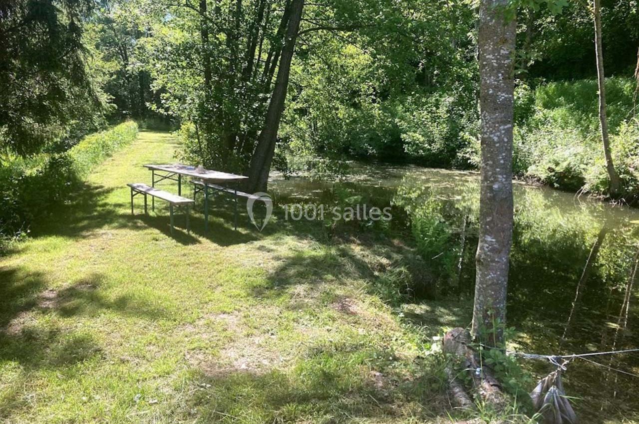 Table de pique-nique en bois près d'un petit cours d'eau entouré de végétation verdoyante sous un ciel ensoleillé.