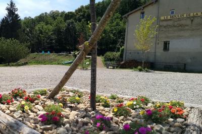 Grande maison en pierre entourée de verdure, avec une cour ensoleillée et des arbres en arrière-plan.