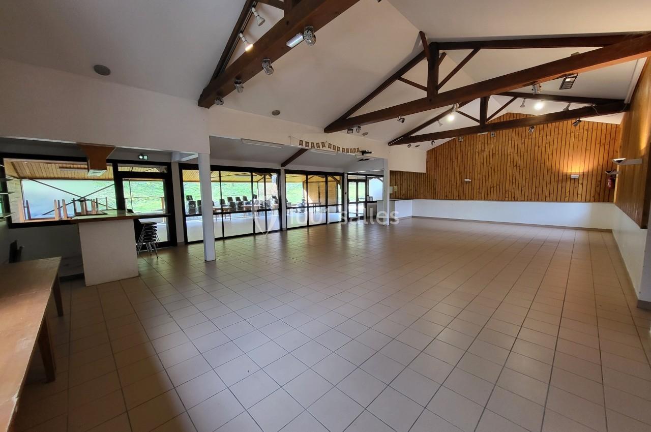 Salle spacieuse avec carrelage au sol, poutres apparentes et grandes baies vitrées donnant sur l'extérieur.