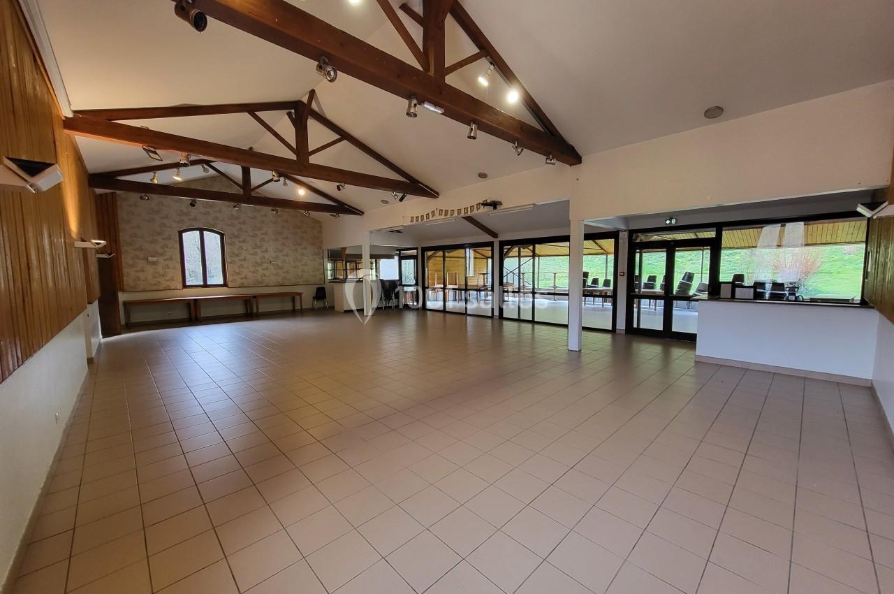 Salle spacieuse avec plafond en poutres apparentes, carrelage au sol et grandes baies vitrées donnant sur l'extérieur.