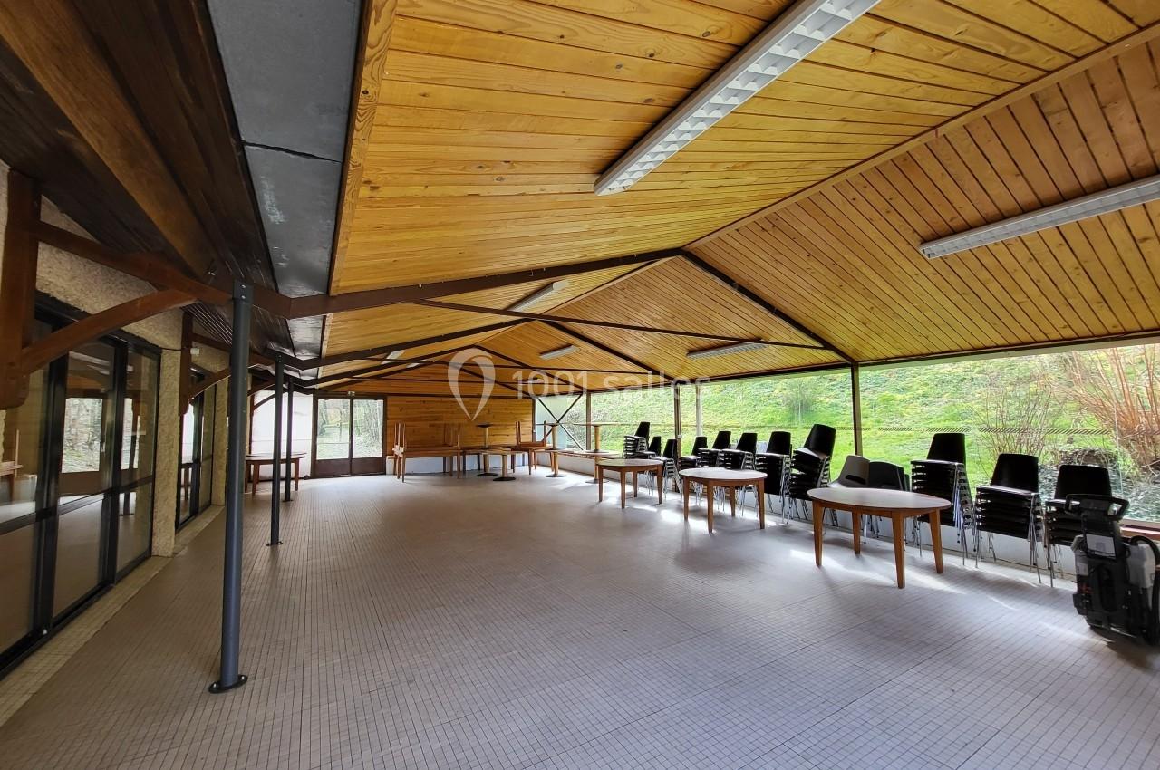 Salle lumineuse avec plafond en bois, tables et chaises empilées près des fenêtres donnant sur un espace verdoyant.