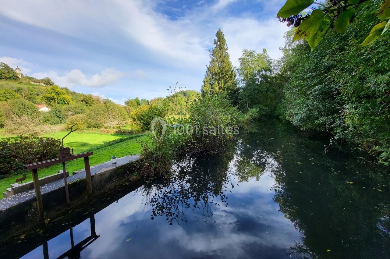 Vue d'un petit cours d'eau entouré de végétation et d'arbres, reflétant un ciel partiellement nuageux.
