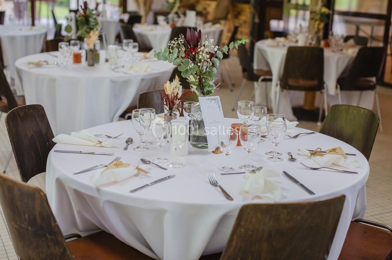 Tables rondes dressées avec nappes blanches, verres, couverts et décorations florales dans une salle de réception.