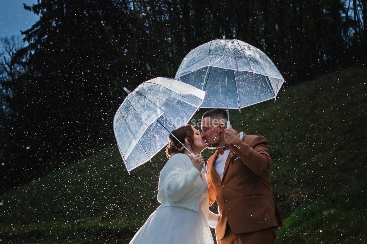 Un couple sous la pluie partage un baiser, abrité par des parapluies transparents dans un paysage naturel en soirée.