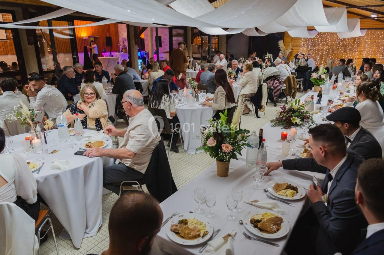 Salle de réception décorée, des invités dînent à des tables rondes avec nappes blanches et arrangements floraux.