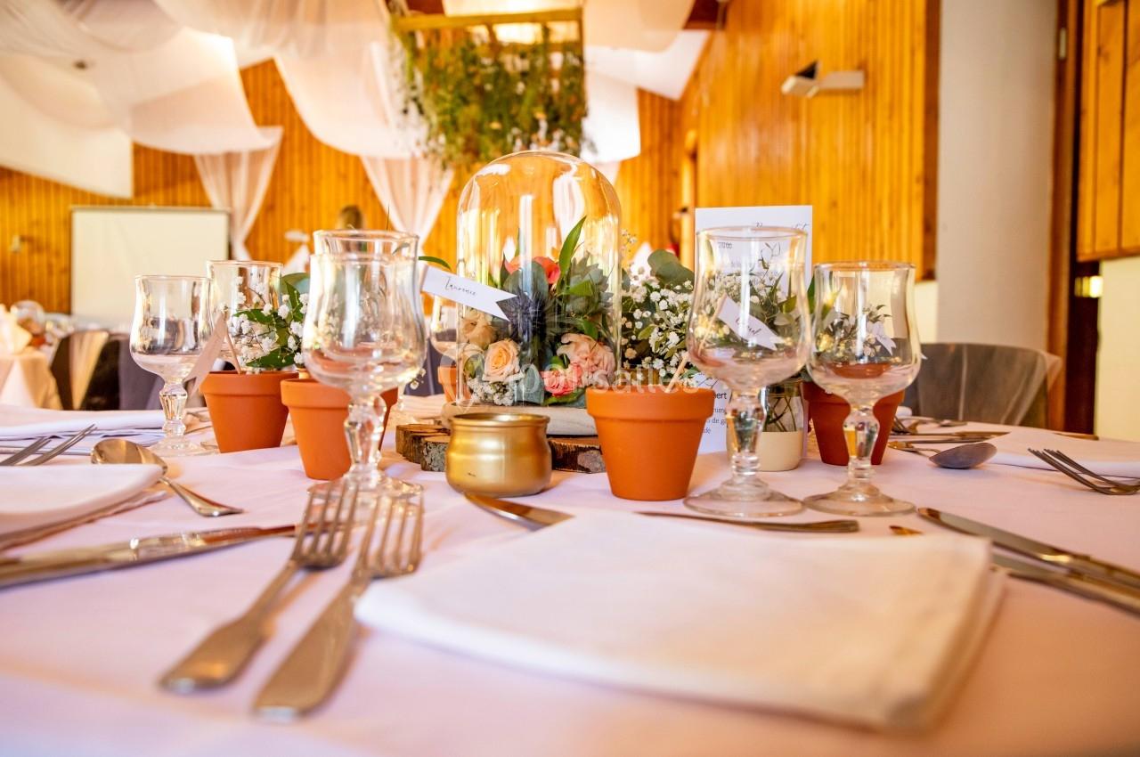 Table décorée pour un événement avec nappes blanches, verres, couverts, plantes en pots et centre de table floral sous…