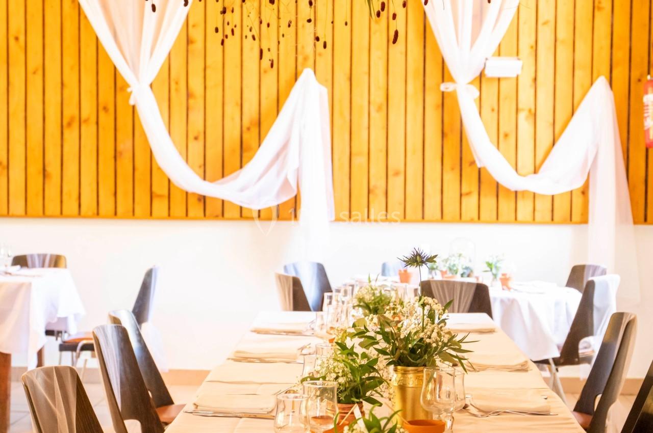 Grande table décorée de plantes et de fleurs, avec nappes blanches, dans une salle lumineuse aux murs en bois.