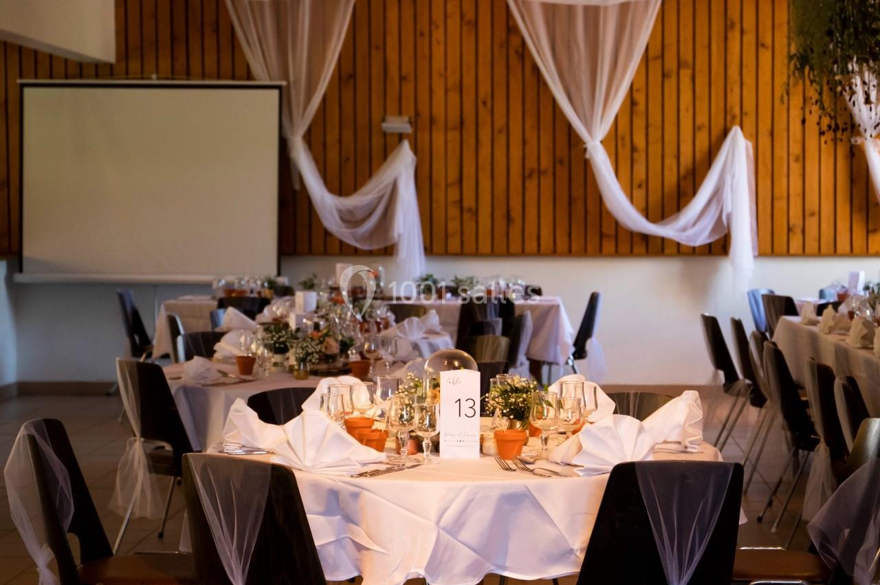 Salle de réception décorée avec des tables rondes, nappes blanches, chaises ornées de voiles et éclairage tamisé.