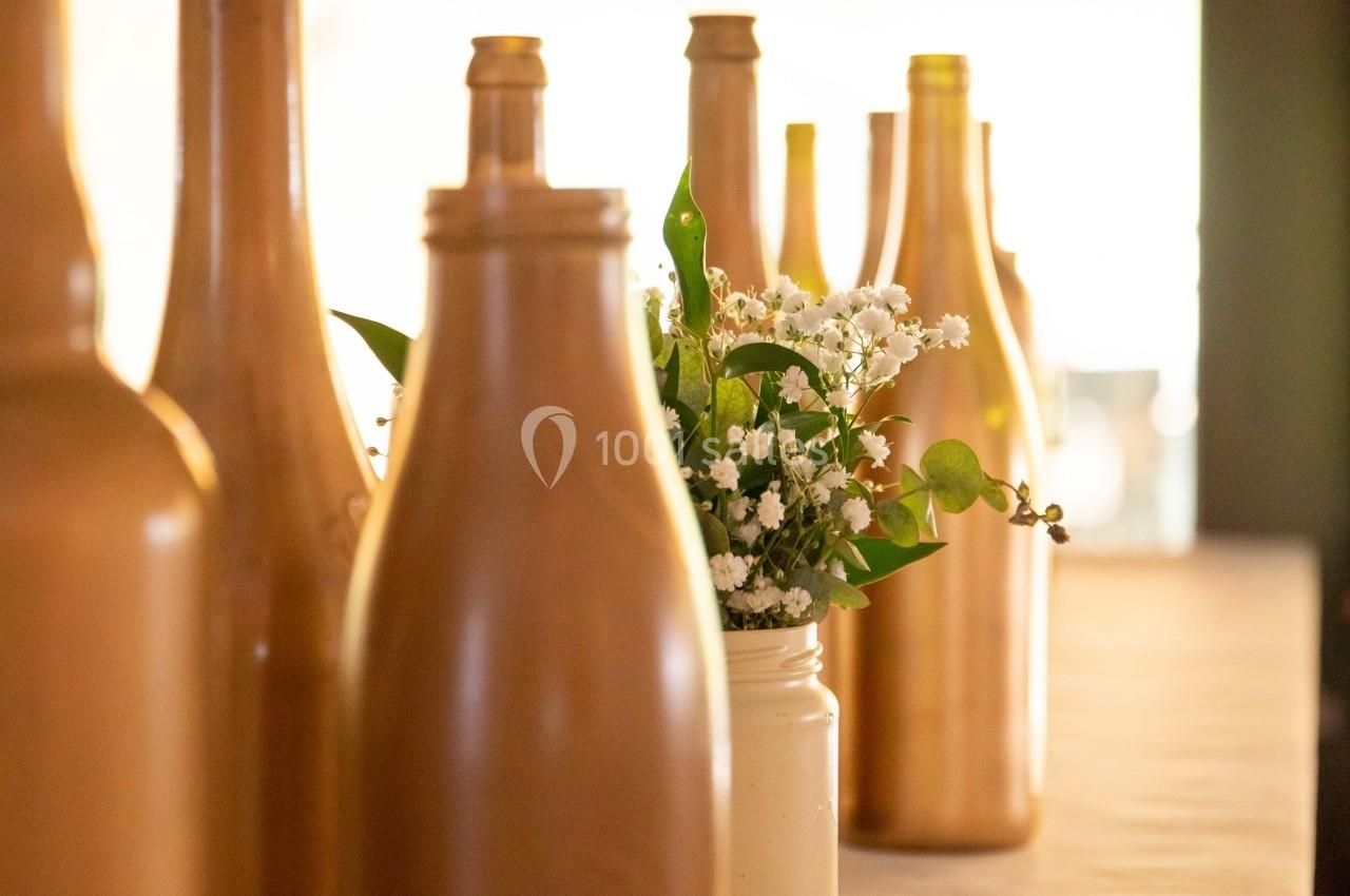 Bouteilles dorées et un vase blanc avec des fleurs blanches disposés sur une table éclairée par une lumière naturelle.